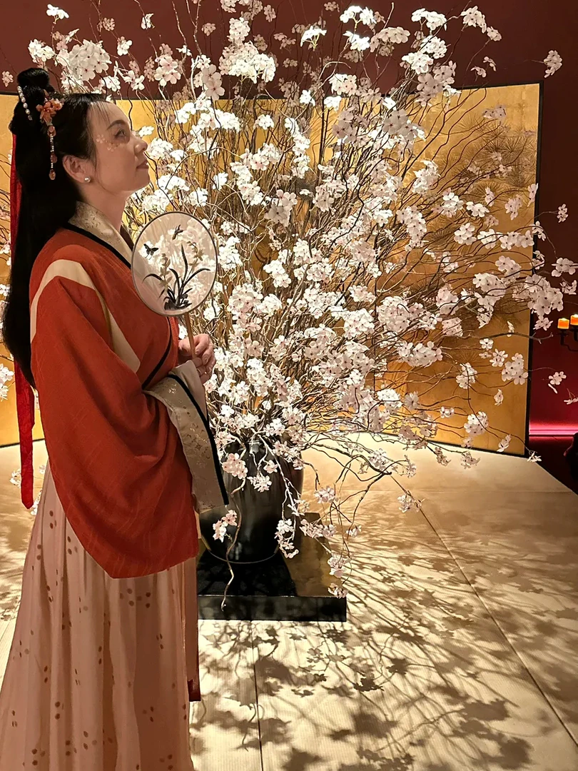 Alicia in a vermilion Hanfu holding a round fan among white blossoms in Shanghai